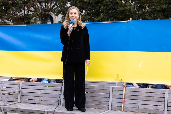 Utomhusbild på Rosencrantz i helfigur med en mikrofon i vänster hand och står på en parkbänk. Bakom henne en banderoll som sträcker sig genom hela bilden. Banderollen har Ukrainas färger.