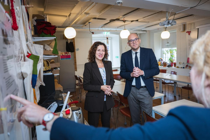 Inomhusbild. Britz och Mînzatu står i ett klassrum vända mot kameran. I förgrunden skymtar en person som pekar mot en text som hänger på väggen till vänster i bild. Skolbänkar och stolar i bakgrunden.