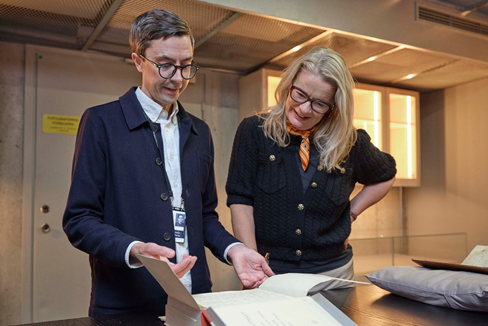 Inomhusbild med Edholm i halvfigur. Hon står bakom ett bord och tittar ned på en bok som visas fram av en person till vänster om henne.