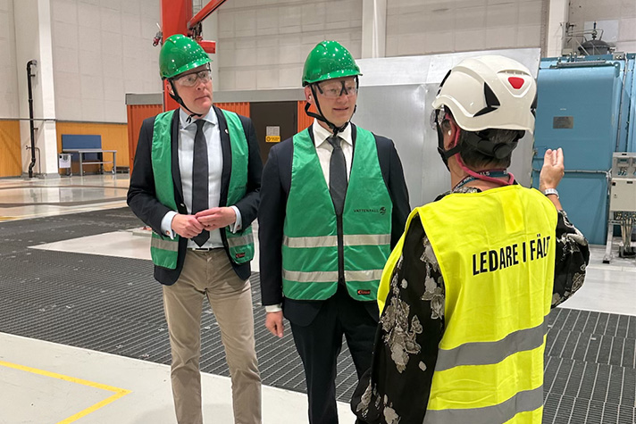 Inomhusbild i modern industrimiljö. Wykman i mitten av bilden bär skyddshjälm, skyddsglasögon och skyddsväst ovanpå kostym. Han samtalar med en person som också bär hjälm och skyddsväst, på ryggen står Ledare i fält. Till vänster om Wykman står ytterligare en person med skyddsutrustning. 