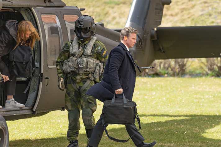 Utomhusbild med Jonsson i profil som går från en helikopter mot höger i bilden. Han bär på en vadderad portfölj. Bakom honom är en person på väg ut ur helikoptern och bredvid öppningen står en militärklädd person med en hjälm som täcker ansiktet och har mörkt tonade glasögon. Helikoptern står på en solig gräsmatta och skuggar de som syns i bild.