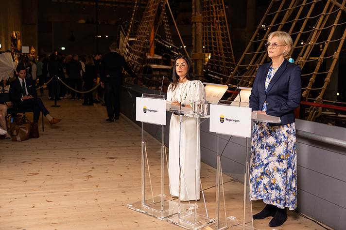 Inomhusbild på Vasamuseet. Liljestrand och Forseke står i strålkastarljus framför Vasaskeppet, vid varsin talpulpet som har en skylt som det står regeringen på. I den mörka bakgrunden syns riggen på skeppet, och man anar en stående publik och sittande journalister.