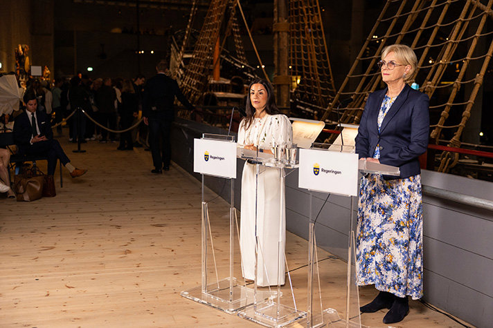 Inomhusbild på Vasamuseet. Liljestrand och Forseke står i strålkastarljus framför Vasaskeppet, vid varsin talpulpet som har en skylt som det står regeringen på. I den mörka bakgrunden syns riggen på skeppet, och man anar en stående publik och sittande journalister.