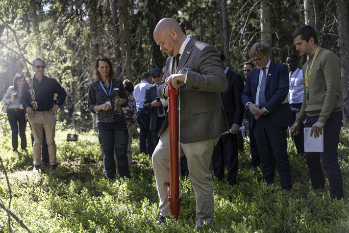 Utomhusbild i en sommarskog med mycket blåbärsris. Kullgren i helfigur står i mitten med ett redskap i händerna. Solen glimtar fram genom grankvistar från vänster i bild. Ett flertal personer med besöksbrickor runt halsen står runt omkring och bakom Kullgren och tittar mot honom.