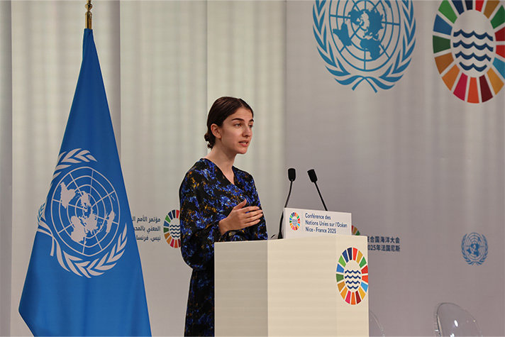 Inomhusbild på Pourmokhtari snett från sidan. Hon står vid en talarstol som har två mikrofoner. Bakom henne står en FN-flagga, och det hänger ett slags tygdraperier i bakgrunden som också har FN-märkning.