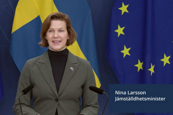 Jämställdhetsminister Nina Larsson står vid en talarpupet. Det är flaggor i fonden.