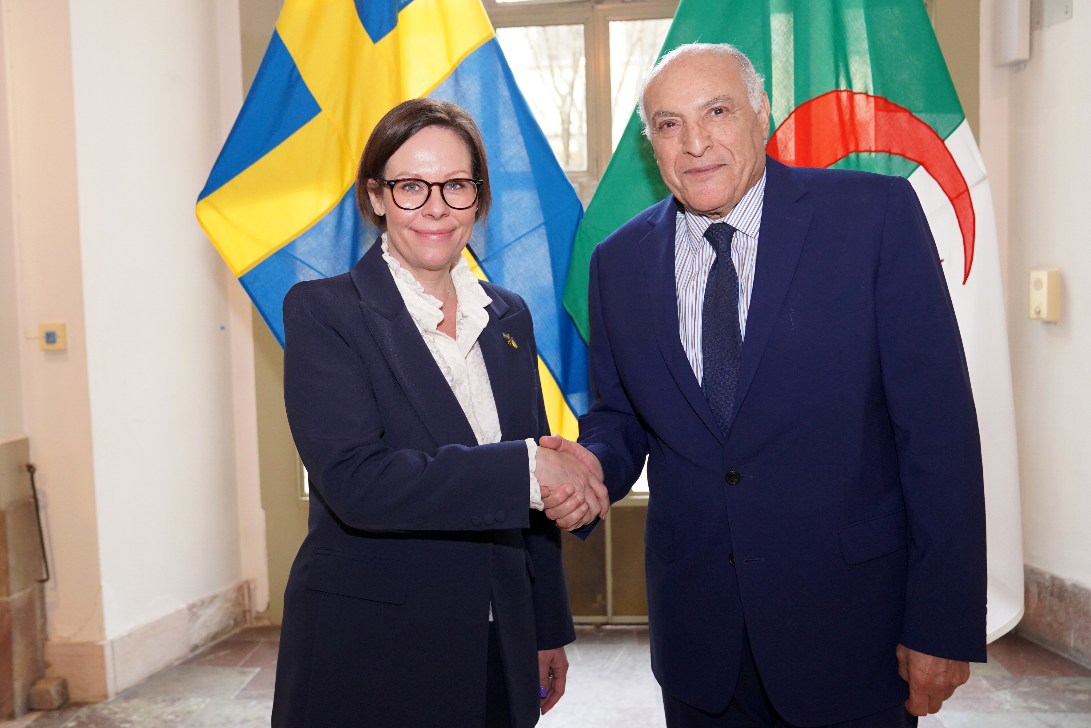 Utrikesminister Maria Malmer Stenergard och Algeriets utrikesminister Ahmed Attaf skakar hand och tittar mot kameran. 
