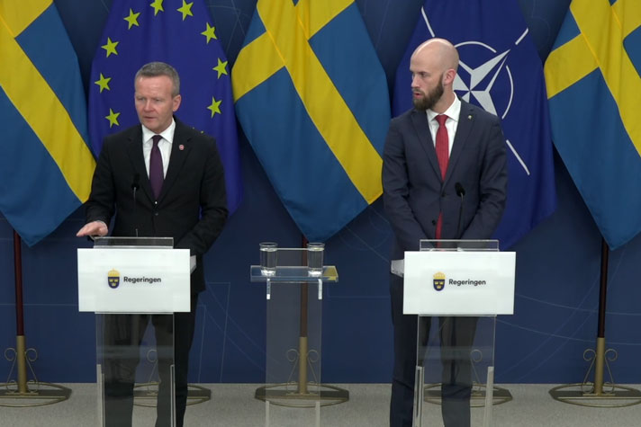 De tv&#xE5; medverkande st&#xE5;r vid var sin talarpulpet. Det &#xE4;r fanor i fonden.