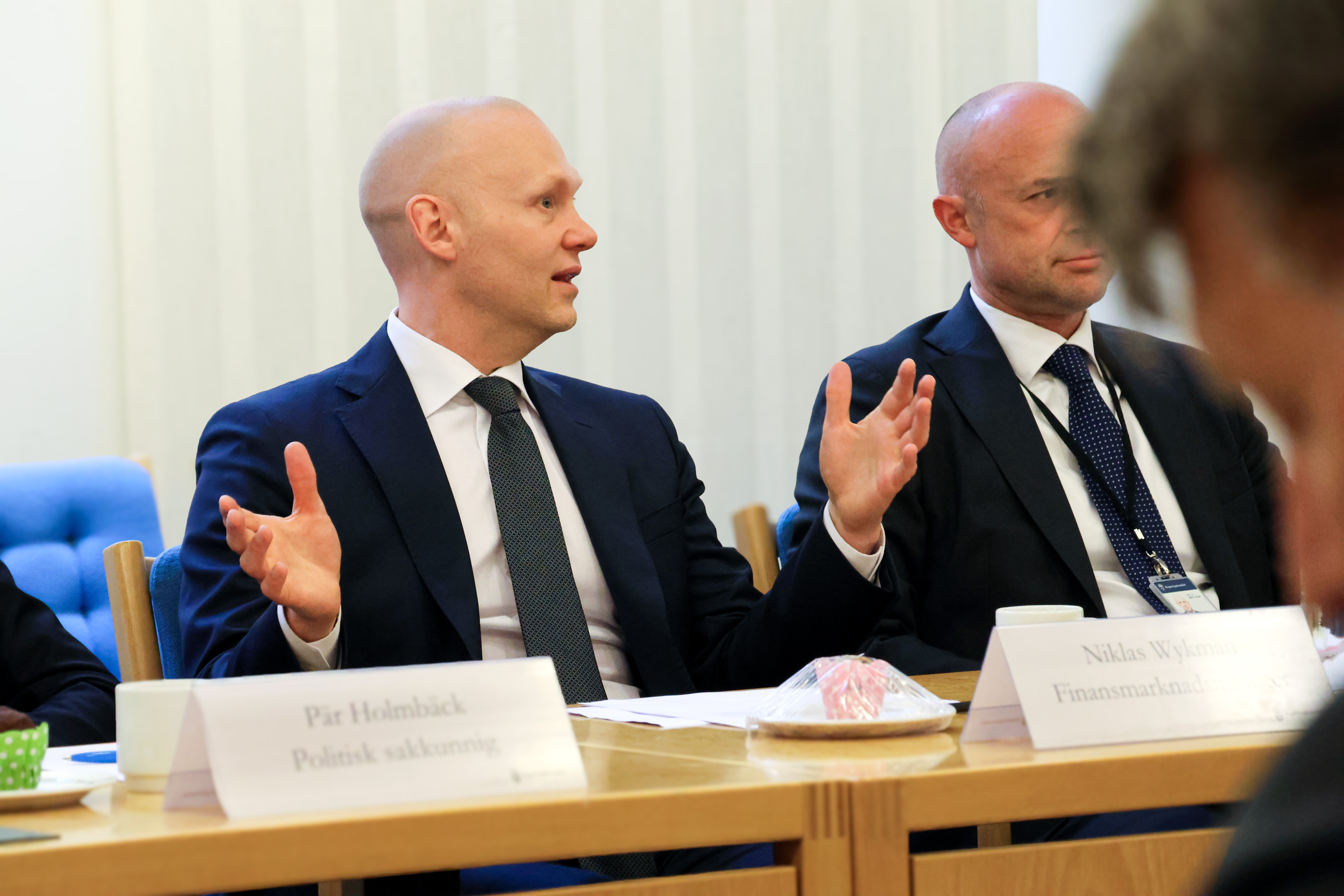 Man sitter vid ett bord och pratar och gestikulerar med händerna. Man anar en ytterligare person bredvid honom, och en annan i förgrunden.