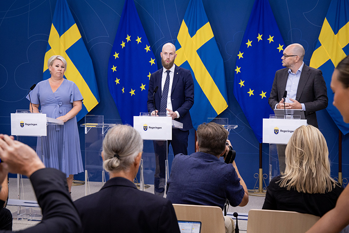 En kvinna och två män står på podiet på en pressträff. I förgrunden finns flera fotografer. I bakgrunden EU-flaggor och svenska flaggor.