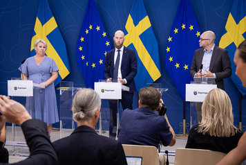 En kvinna och två män står på podiet på en pressträff. I förgrunden finns flera fotografer. I bakgrunden EU-flaggor och svenska flaggor.