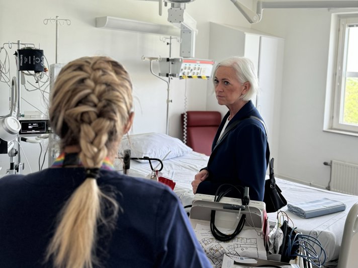 Sjukvårdsminister Acko Ankarberg Johansson besöker ett patientrum på Visby lasarett. I förgrunden står person i sjukhuskläder med ryggen vänd mot kameran och förklarar något för sjukvårdsministern som lyssnar.