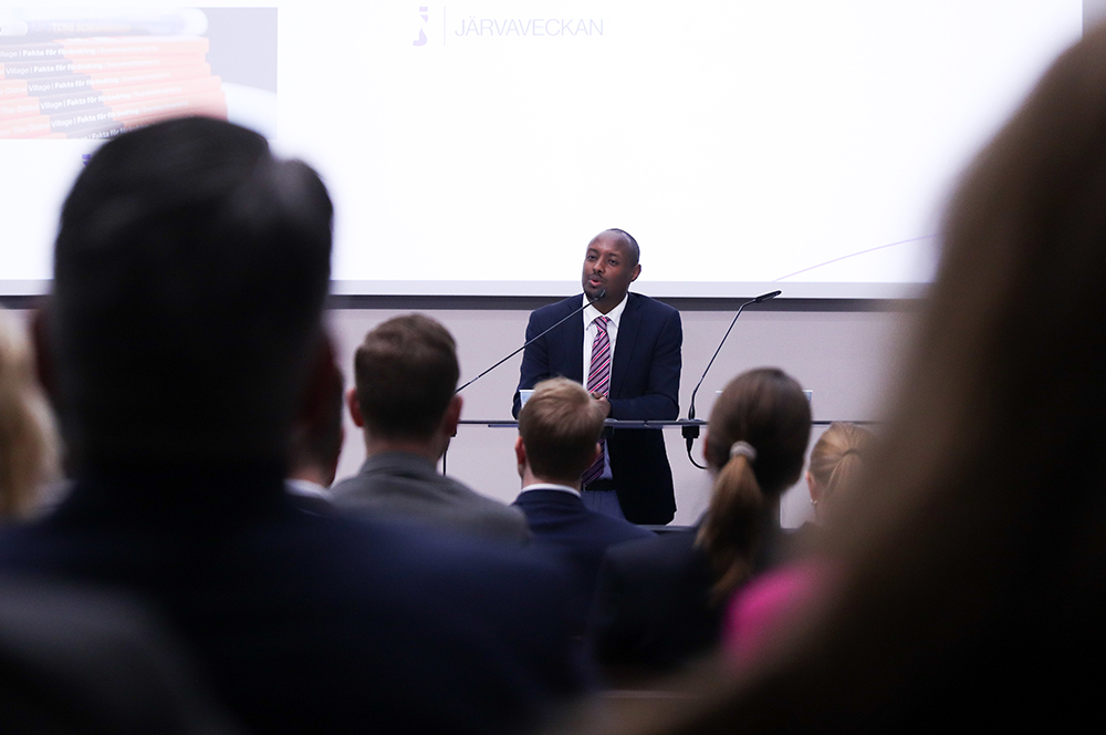 En man talar på en scen. I förgrunden syns personer som sitter i publiken och lyssnar.