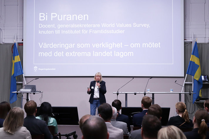 En kvinna talar i en mikrofon på en scen. I förgrunden syns huvuden på publiken. I bakgrunden syns en stor skärm med texten Bi Puranen, Värderingar som verklighet.