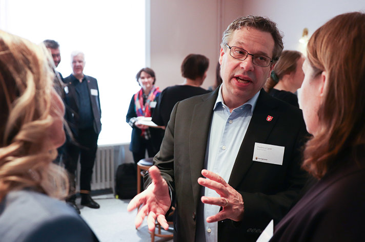 En man gestikulerar med händerna och talar med en kvinna. I bakgrunden syns några andra personer.