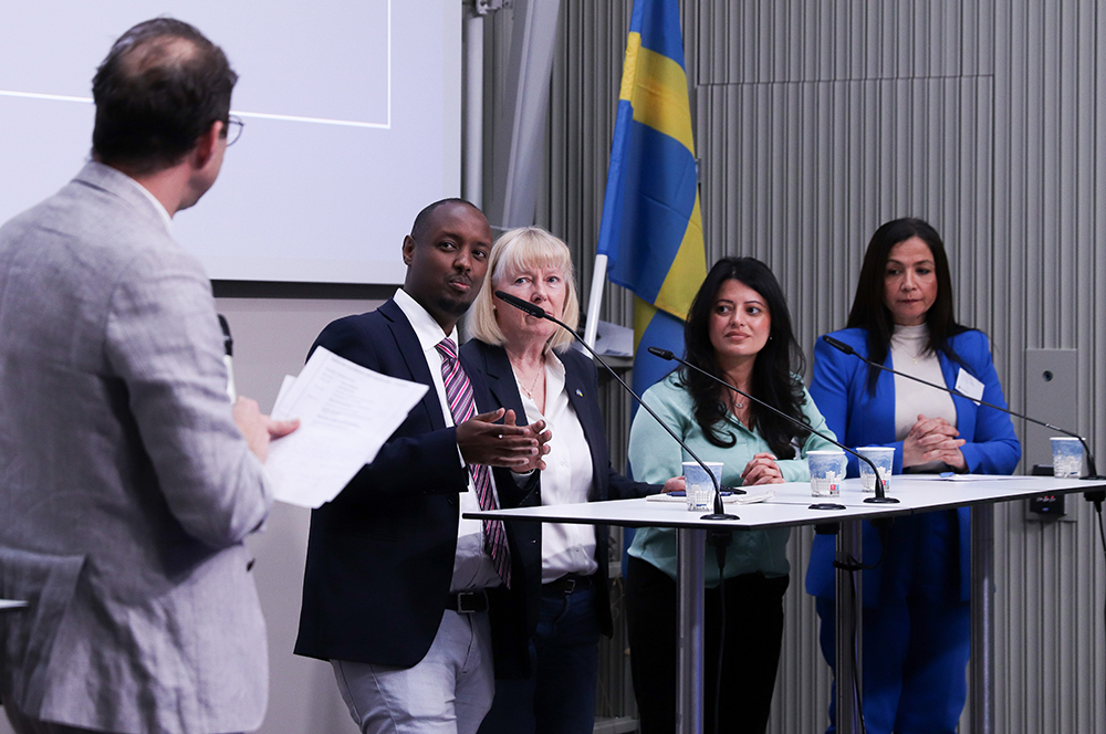 Fyra personer står vid ståbord på en scen. En moderator står med ryggen mot kameran och tittar på panelen. 