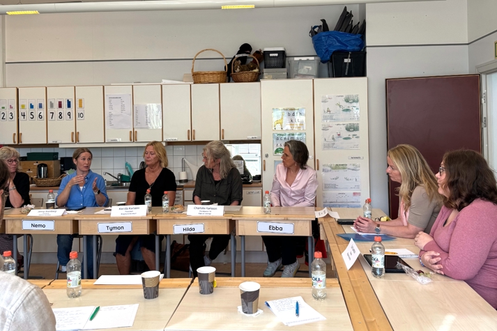 Lärarna och skolministern sitter vid skolbänkar i ett klassrum och diskuterar.