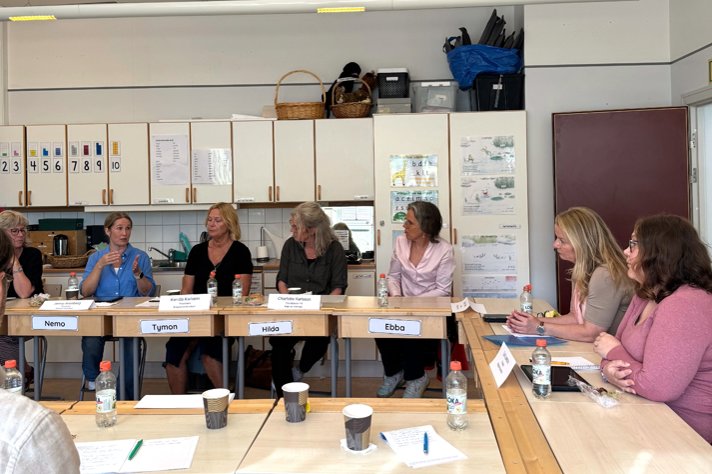 Lärarna och skolministern sitter vid skolbänkar i ett klassrum och diskuterar.