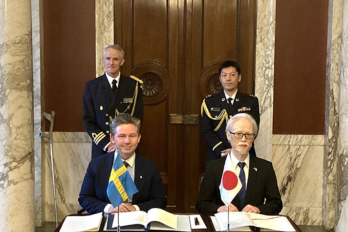 Försvarsminister Pål Jonson och Japans ambassadör i Stockholm H.E. Noke Masaki sitter bredvid varandra vid ett bord. Bakom dem står två män i uniform. På bordet ligger papper och det står även en svensk flagga och en japansk flagga på bordet.