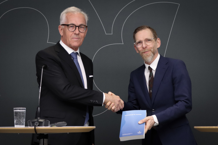 Jan Albert och Jakob Forssmed skakar hand. Forssmed håller upp betänkandet i handen, som Albert lämnat över.