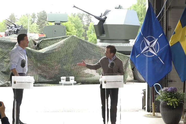 De båda medverkande står vid var sitt ståbord i öppningen till en större byggnad. I fonden utanför skymtar militära fordon och materiel.