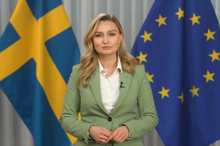 Ebba Busch inomhus framför svenska flaggan och EU-flaggan.