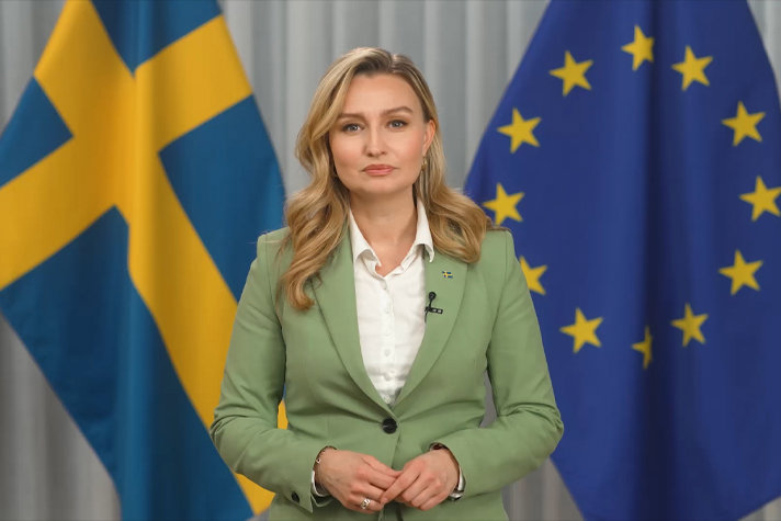 Ebba Busch inomhus framför svenska flaggan och EU-flaggan.