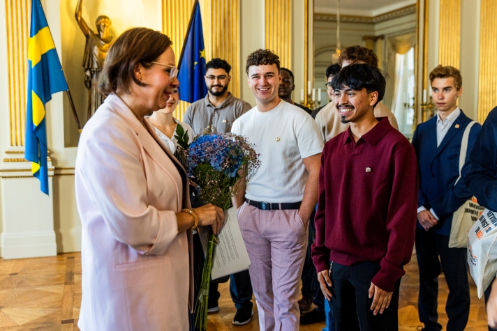 En ung man ger blommor till ministern. En grupp unga människor står bredvid. I bakgrunden skymtar två flaggor, en för Sverige och en för EU.