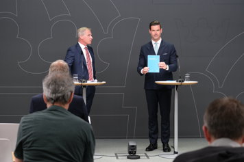 Utredare Ola Sjöstrand och migrationsminister Johan Forssell står framför journalisterna på en pressträff.