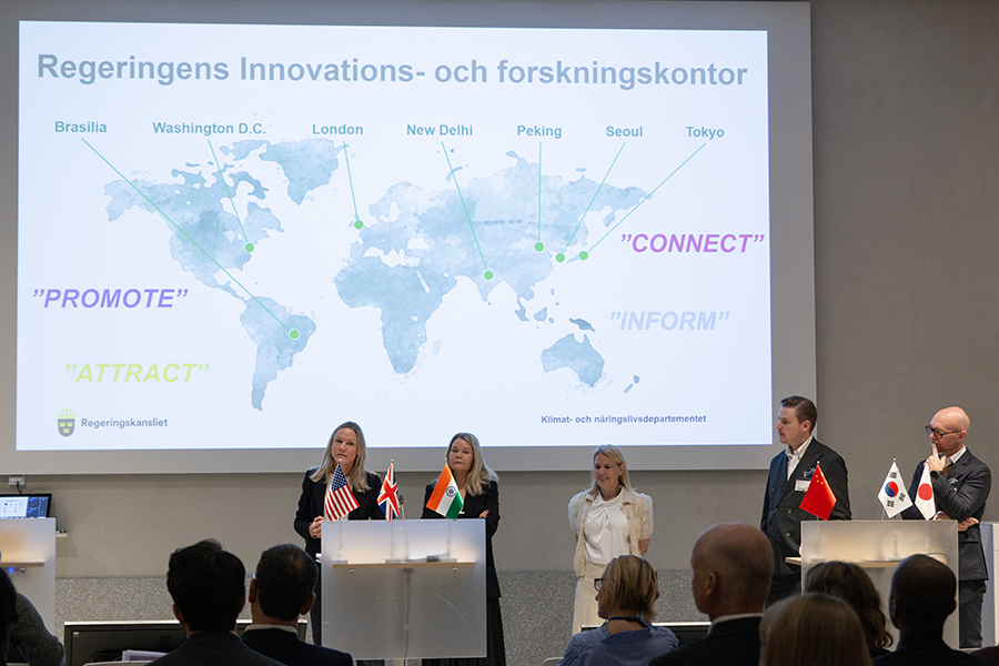 Fem personer som presenterar vad  de som regeringens innovations- och forskningsråd arbetar med. De står i en stor aula framför en en världskarta där de olika kontoren är utmärkta.
