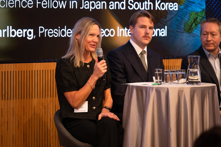 En kvinna talar i en mikrofon, och vid hennes sida sitter två män i en soffa.