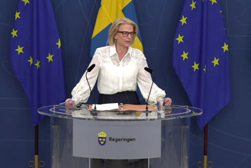 Finansminister Elisabeth Svantesson står vid en talarpulpet med flaggor i fonden.