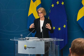 Elisabeth Svantesson står i en talarstol i Regeringskansliets presskonferenssal.  I bakgrunden svenska flaggor och EU-flaggan.