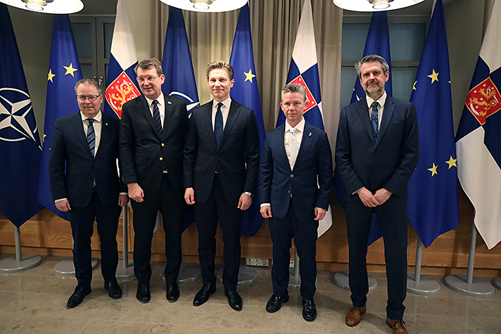 Fem personer står på rad inomhus. I bakgrunden syns olika flaggor. 