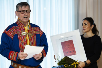 Kulturminister Parisa Liljestrand håller i ett diplom och en blombukett, pristagaren Ole Henrik Magga håller tal.