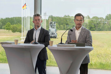 Försvarsminister Pål Jonson och statsminister Ulf Kristersson står vid var sin talarpulpet med ett öppet fält i bakgrunden. På fältet står ett militärfordon.