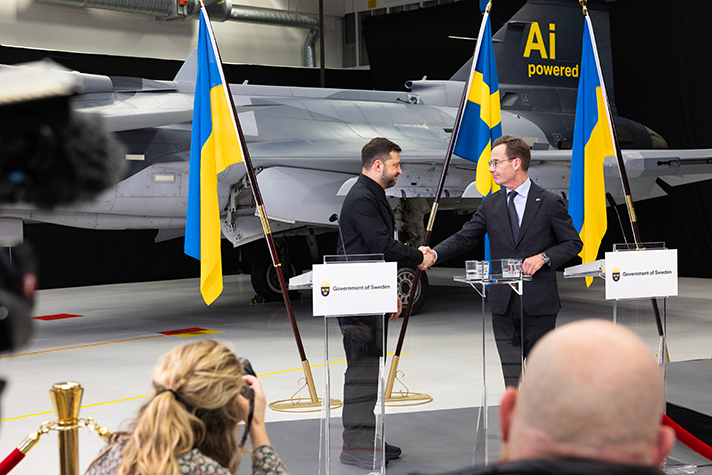 Volodymyr Zelensky och Ulf Kristersson står på en pressträff och skakar hand. I bakgrunden står ett JAS Gripen samt ukrainska och svenska flaggor. I förgrunden syns journalister.