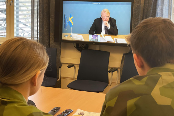 Två unga värnpliktiga sedda bakifrån med en skärm med försvarsministern framför sig.