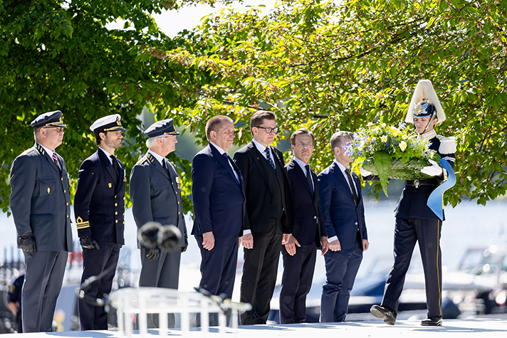 Statsråd, H.M. konungen och överbefälhavaren m.fl. står på rad på ett podium bredvid soldater som håller kransar.