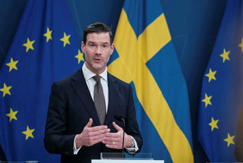 Migrationsminister Johan Forssell står vid en talarpulpet. Flaggor i fonden.