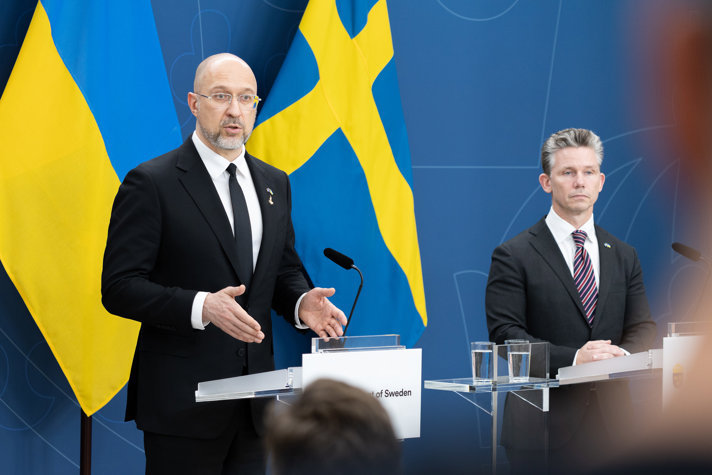 Två män i kostym står vid varsin talarstol och håller pressträff. I bakgrunden skymtar svenska och ukrainska flaggor.