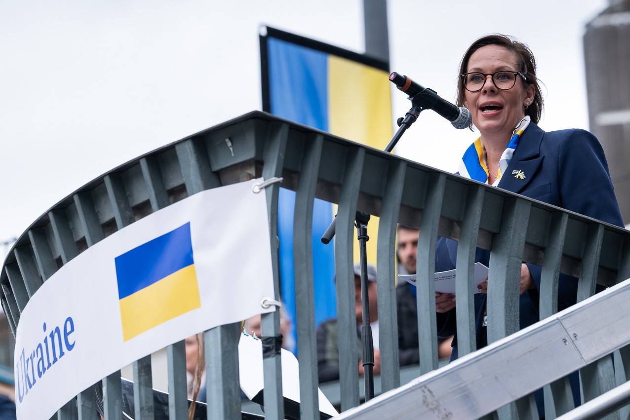 Kvinna talar i mikrofon, ukrainska flaggor i bakgrunden.