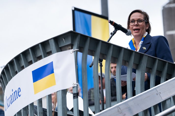 Kvinna talar i mikrofon, ukrainska flaggor i bakgrunden.