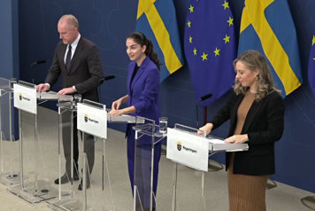 Tre personer står vid var sin talarstol, det är fanor i fonden.