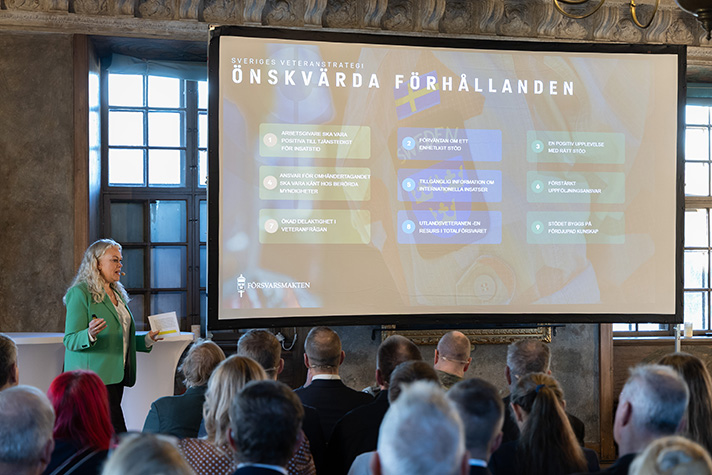 En moderator står vid en overhead display som visar önskvärda förhållanden genom Sveriges veteranstrategi.