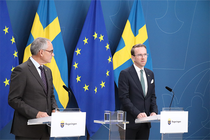 två män i kostym står framför en blå bakgrund med flaggor