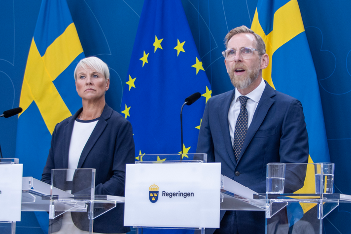 Anna Tenje och Jakob Forssmed på pressträff. Svenska flaggor och EU-flaggor bakom dem.