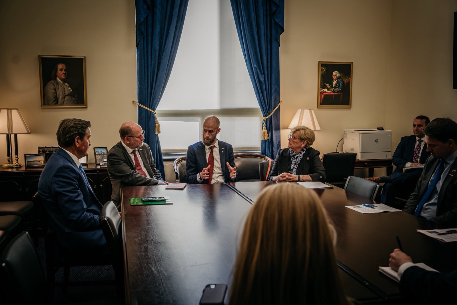 Statsrådet sitter vid ett bord inomhus omgiven av människor.