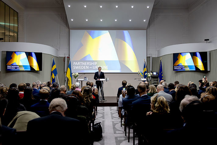 En man håller tal på scenen i en fullsatt konferenslokal. På skärmen bakom honom syns den svenska flaggan och den ukrainska flaggan, och texten Partnership Sweden-Ukraine.