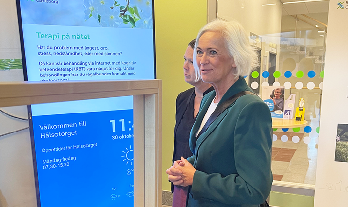 Acko Ankarberg Johansson står vid en skylt där det står "Välkommen till hälsotorget#